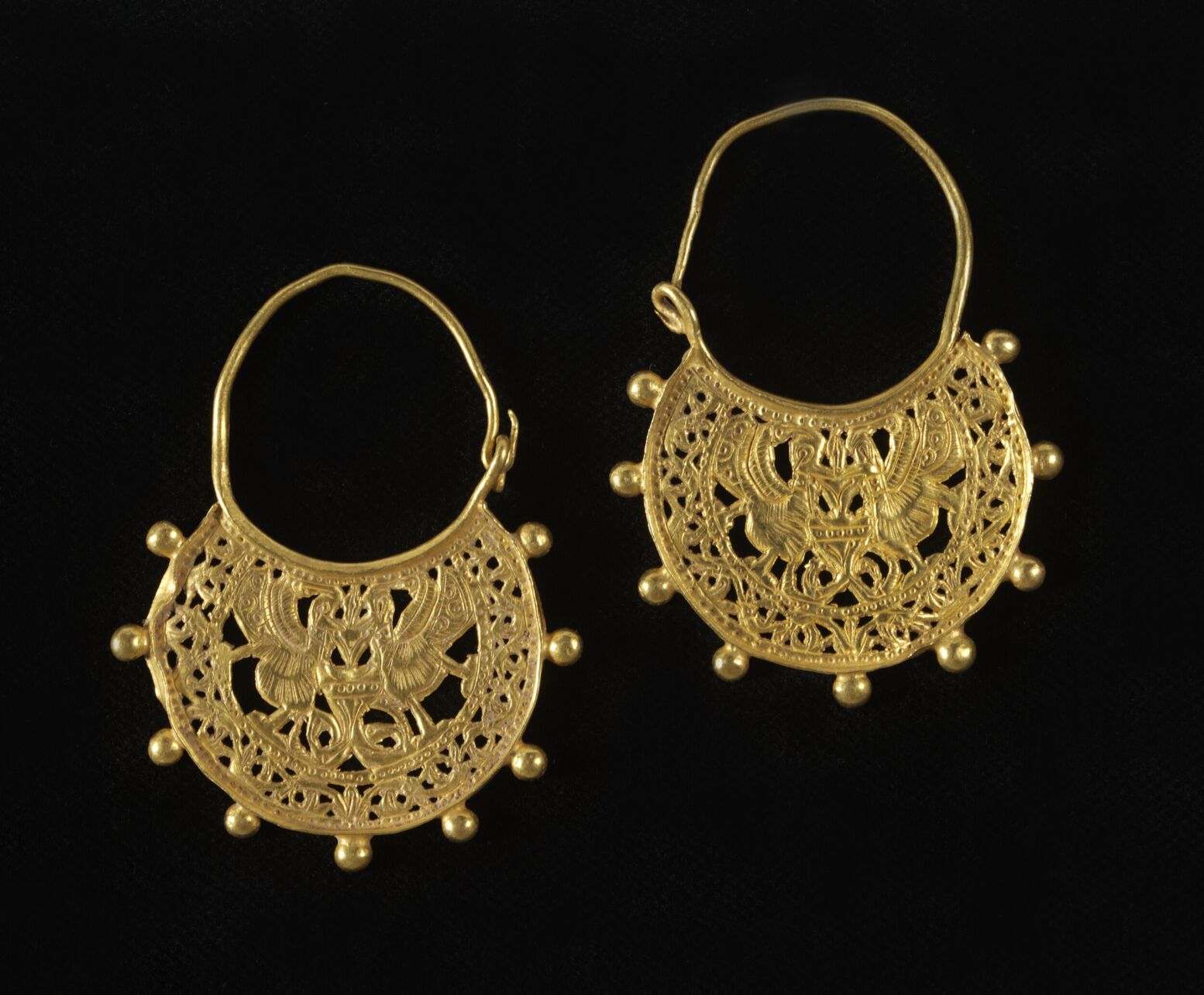 Pair of seventh-century Byzantine crescent-shaped gold earrings with pierced decoration showing peacocks drinking from a well, against a black background.