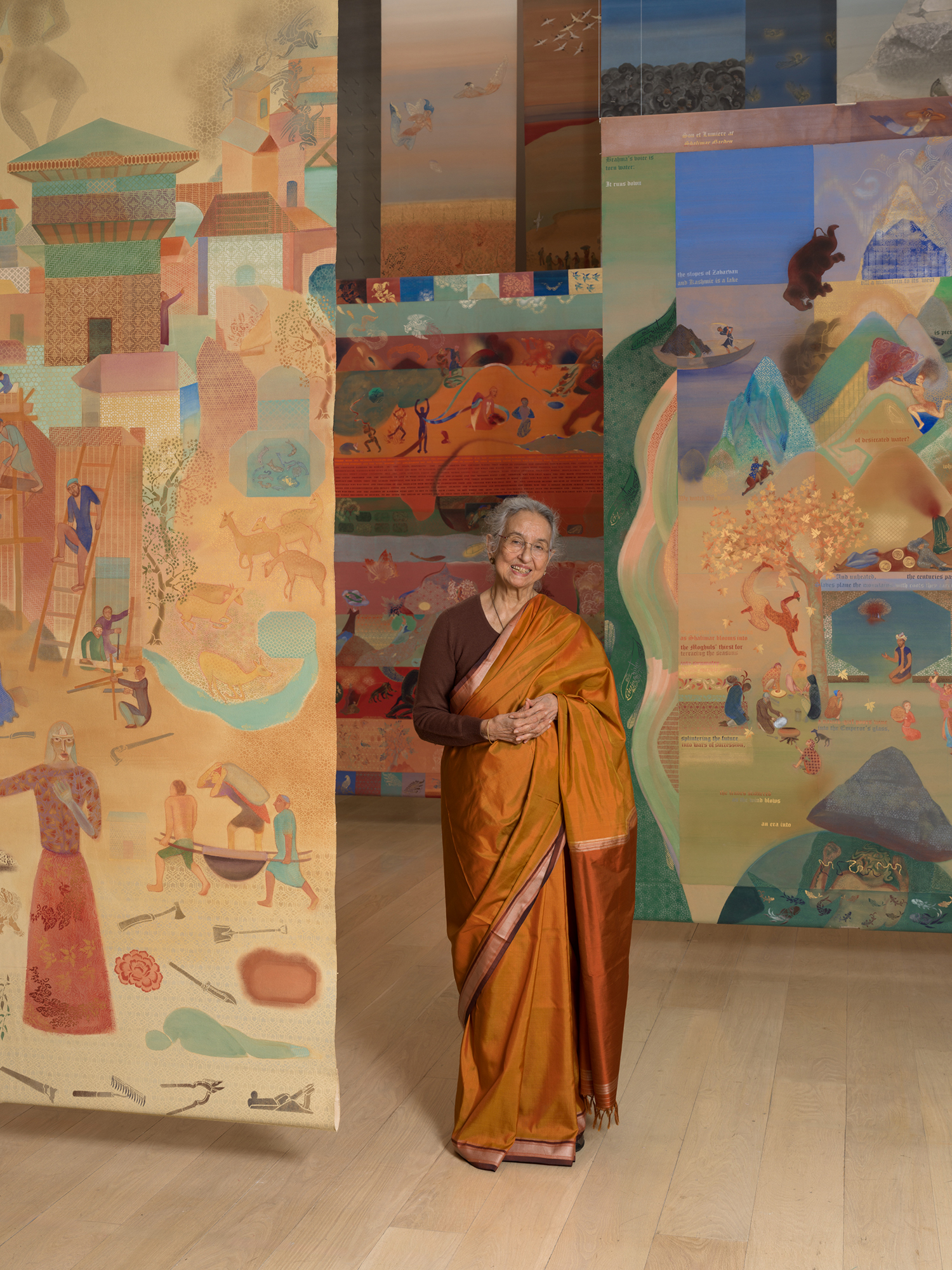 Nilima Sheikh wearing an orange sari, standing among her suspended painted scrolls in Fractured Skies at MoMA.